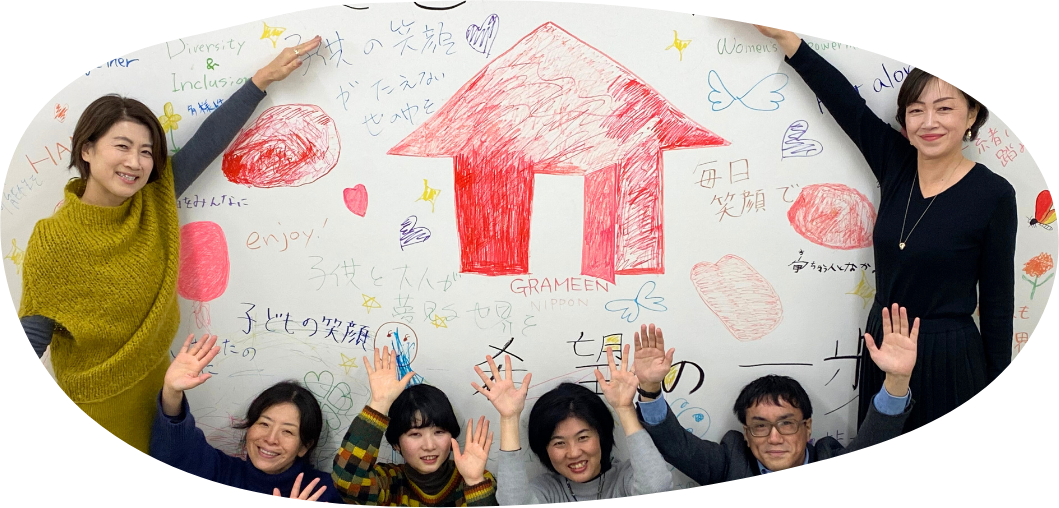 グラミン日本参加者の集合写真。背景にグラミン日本のロゴの家があり、6名の男女が手を挙げて映っている。